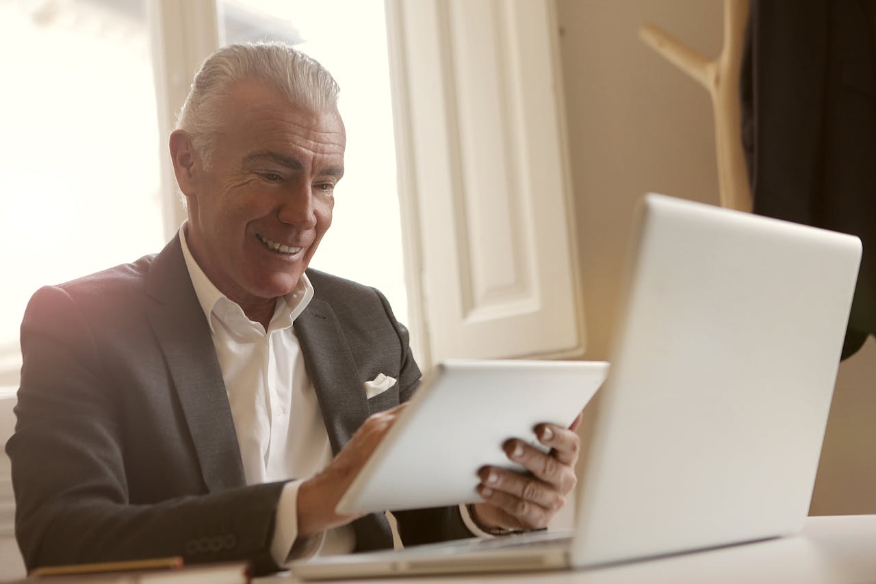 Smiling senior man multitasking with a laptop and tablet indoors at a desk.
