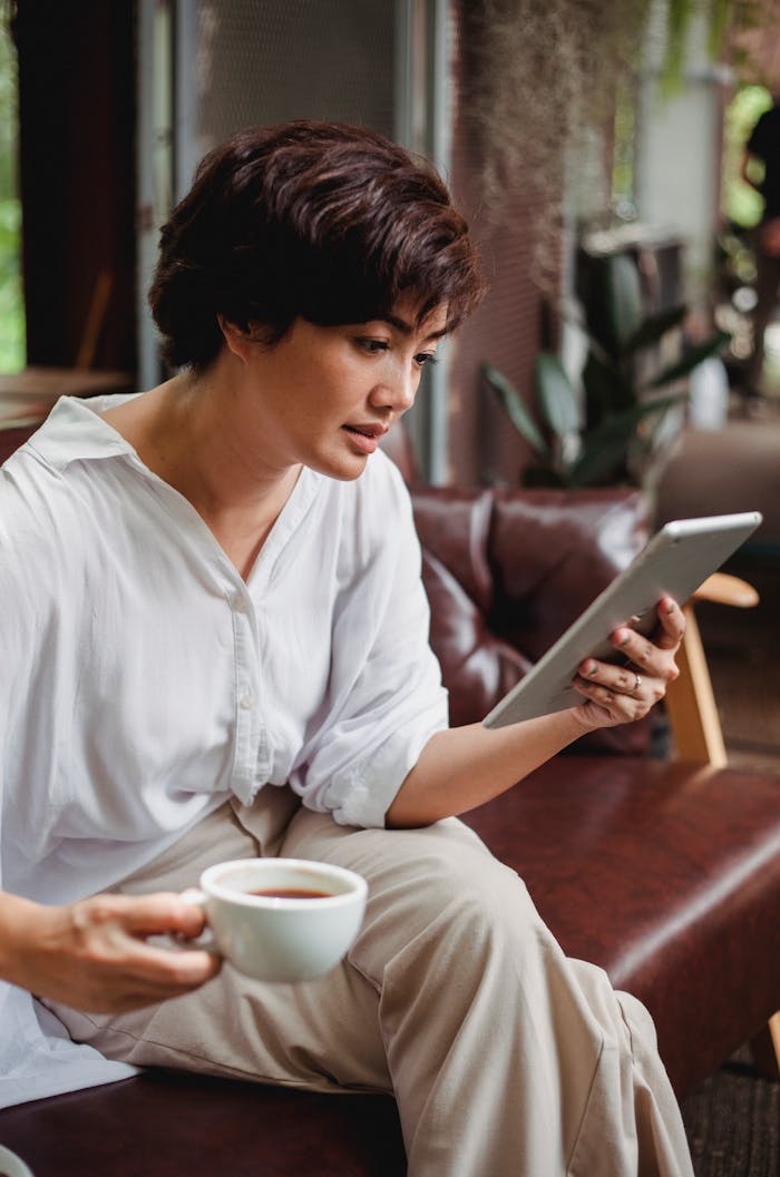 Side view of concentrated Asian woman in stylish clothes raising hand with cup of hot drink and browsing tablet in daytime