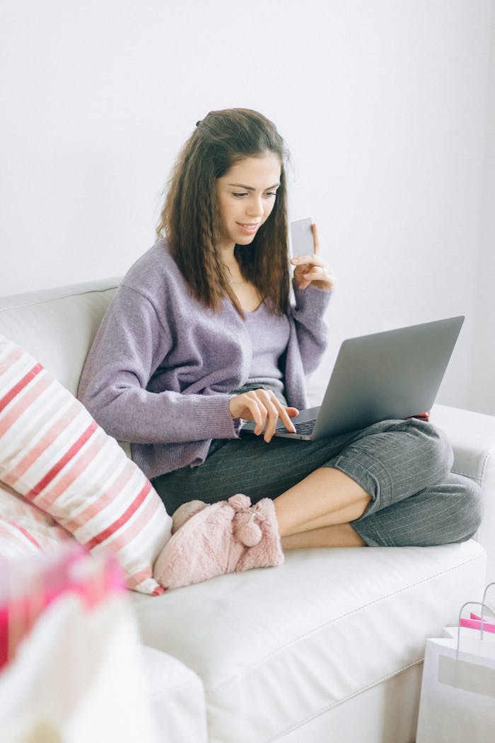 Woman in cozy attire shopping online with laptop and credit card on the couch.