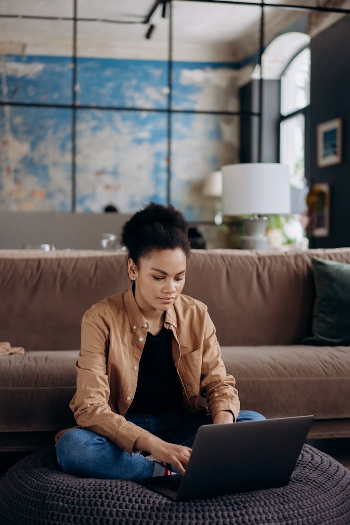 A woman sits on a floor cushion, working on a laptop in a modern living room.